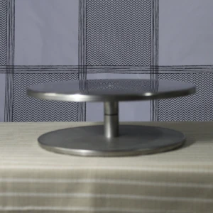 Soporte giratorio de acero inoxidable con base y plato circular, sobre mesa con tela a rayas, ideal para decoración de pasteles.