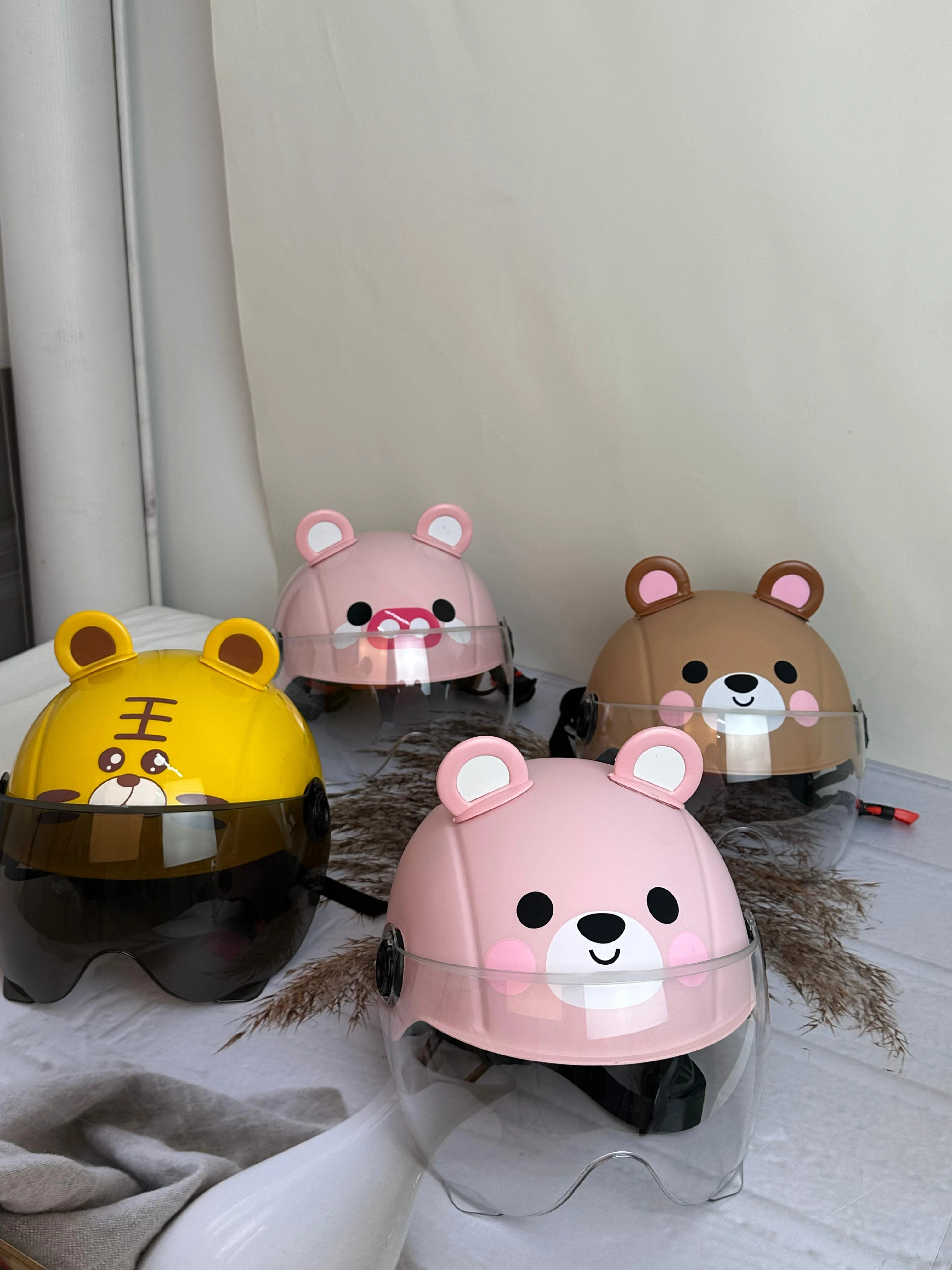 Cuatro cascos infantiles de diseño animal con visor transparente y ajuste seguro, en colores rosa, amarillo y marrón, dispuestos sobre tela clara.