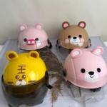 Cuatro cascos infantiles con visor transparente y diseño de animales: cerdo rosa, oso marrón, tigre amarillo y oso rosa, dispuestos sobre tela blanca con pluma decorativa.
