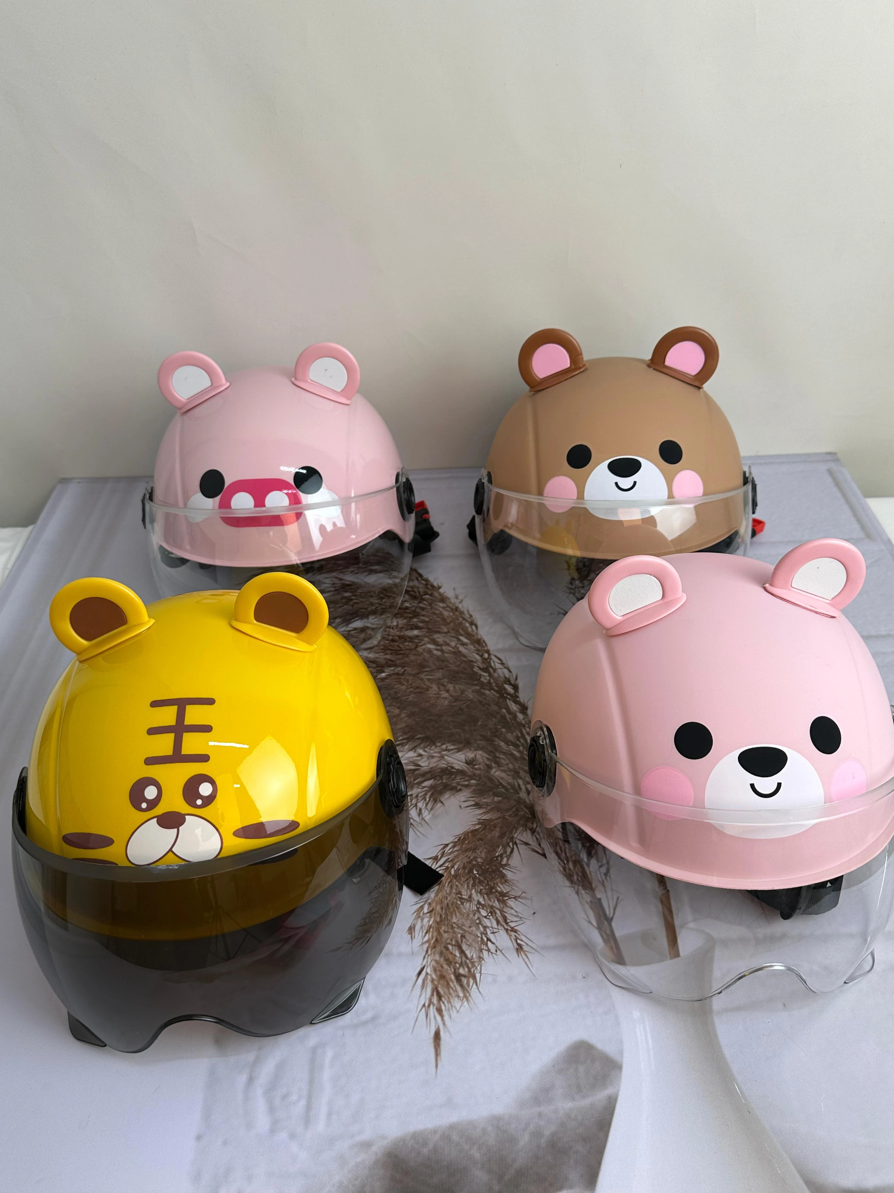 Cuatro cascos infantiles con visor transparente y diseño de animales: cerdo rosa, oso marrón, tigre amarillo y oso rosa, dispuestos sobre tela blanca con pluma decorativa.
