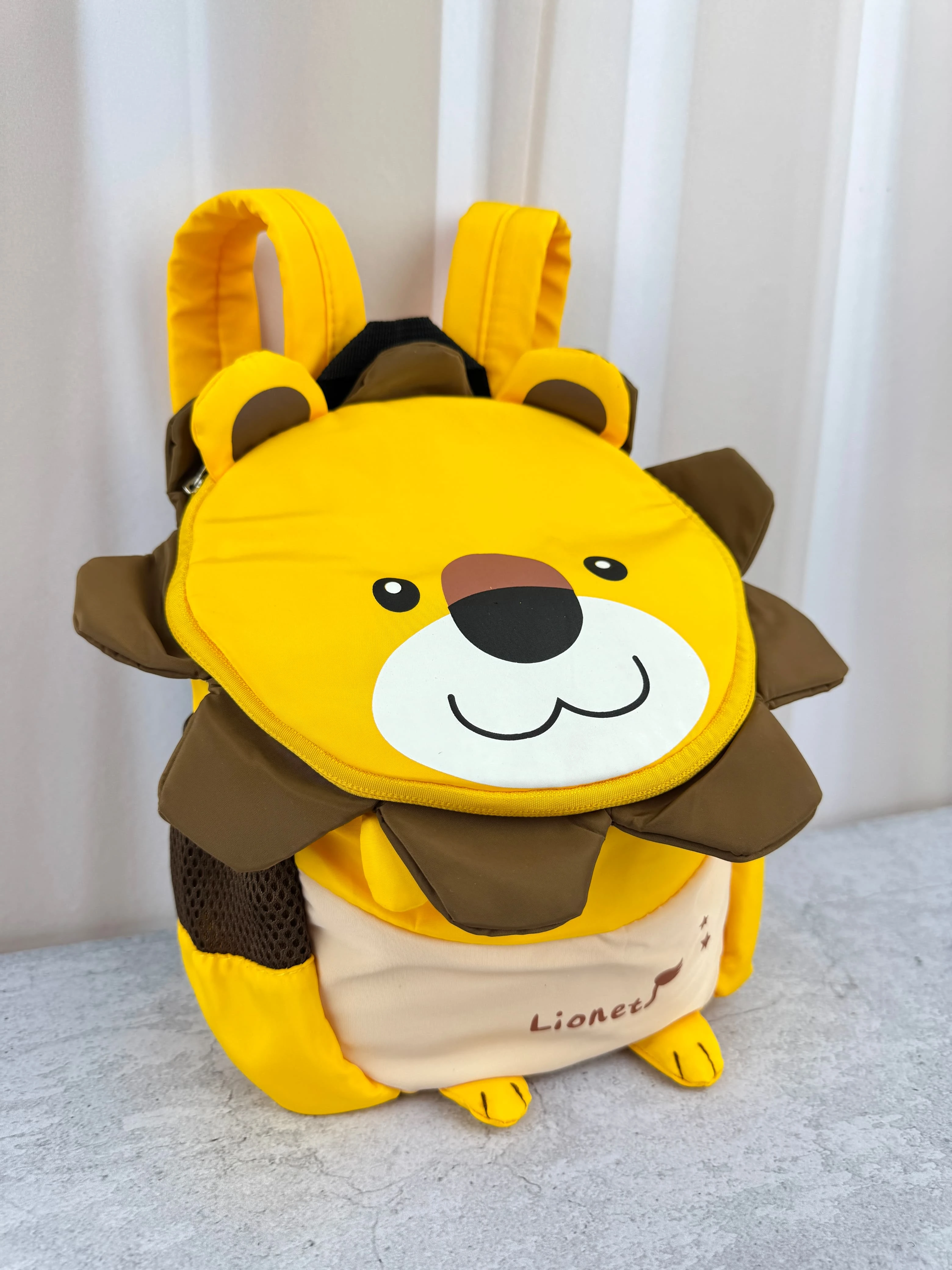 Mochila infantil amarilla con diseño de león, incluye correas de seguridad y detalles en marrón y beige.