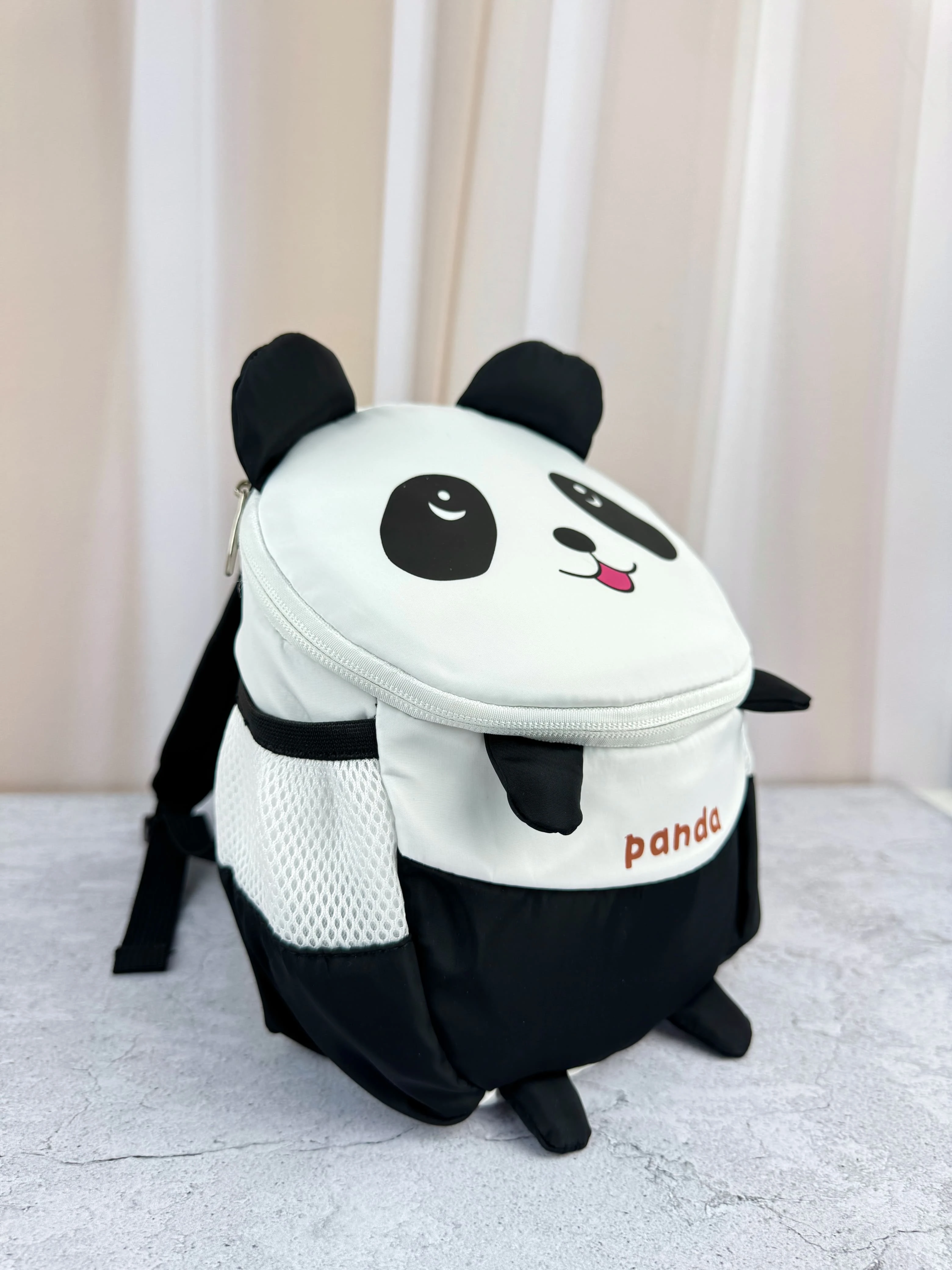 Mochila infantil panda con diseño 3D, color blanco y negro, malla lateral y correas de seguridad visibles.