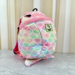 Mochila infantil con estampado de escamas multicolor, correas rosas y parche verde en la parte frontal, sobre superficie gris.