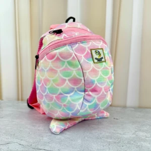 Mochila infantil con estampado de escamas multicolor, correas rosas y parche verde en la parte frontal, sobre superficie gris.