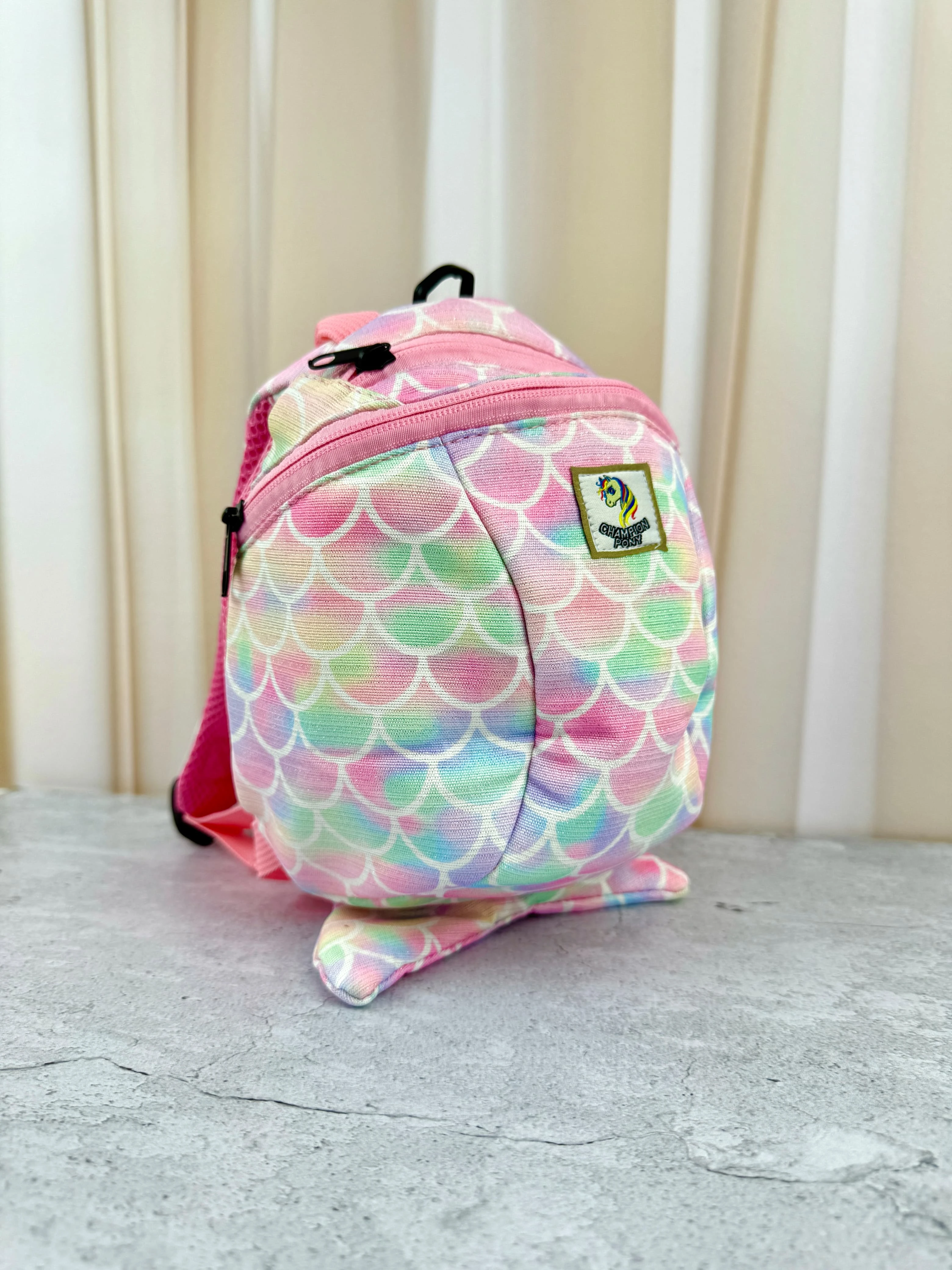 Mochila infantil con estampado de escamas multicolor, correas rosas y parche verde en la parte frontal, sobre superficie gris.