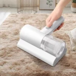 Aspiradora de mano portátil blanca con filtro HEPA, en uso sobre alfombra beige, eliminando ácaros y alérgenos de sábanas y tapicería.