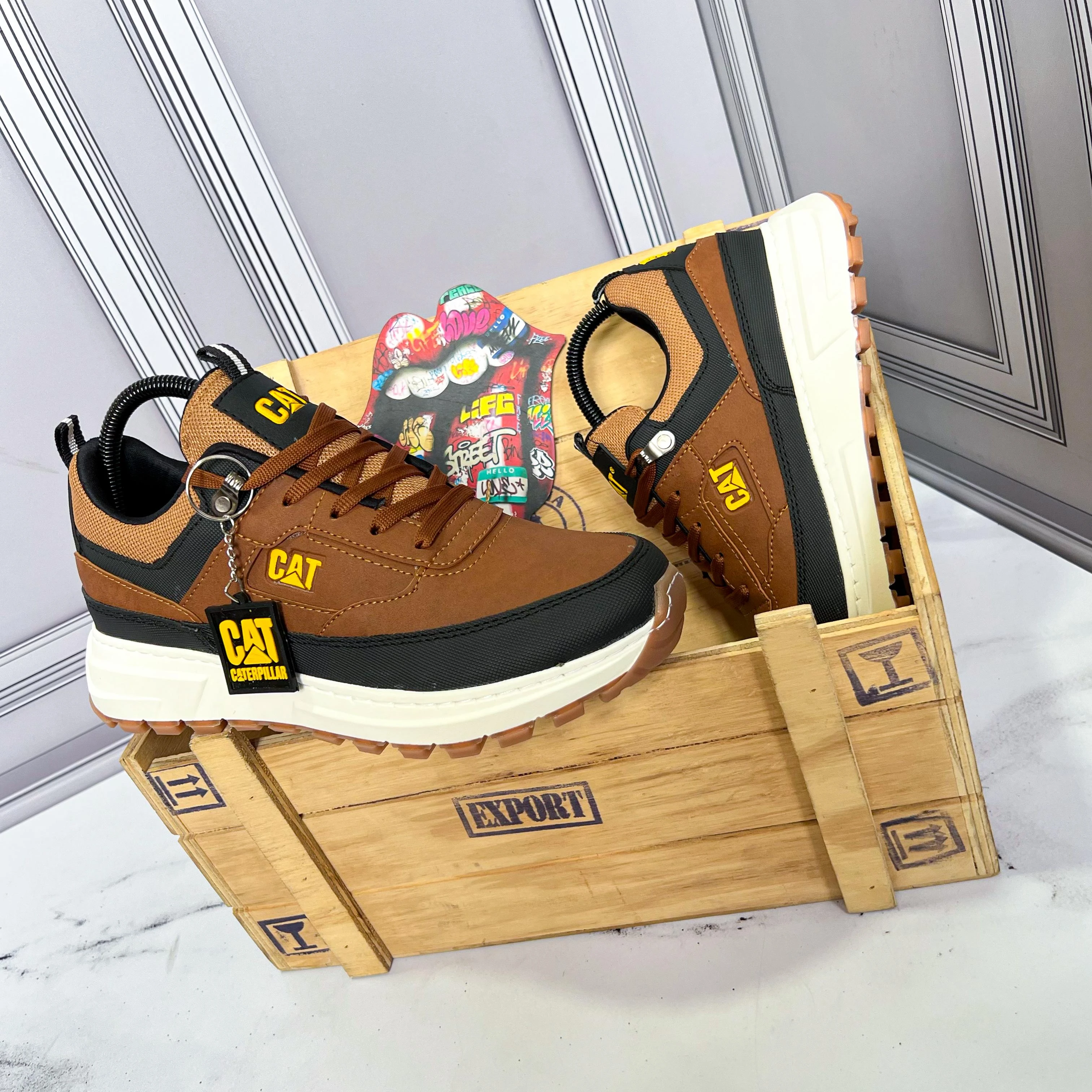Zapatos deportivos sintéticos Caterpillar en tono marrón con detalles negros y suela blanca, presentados sobre caja de madera con etiqueta colgante.