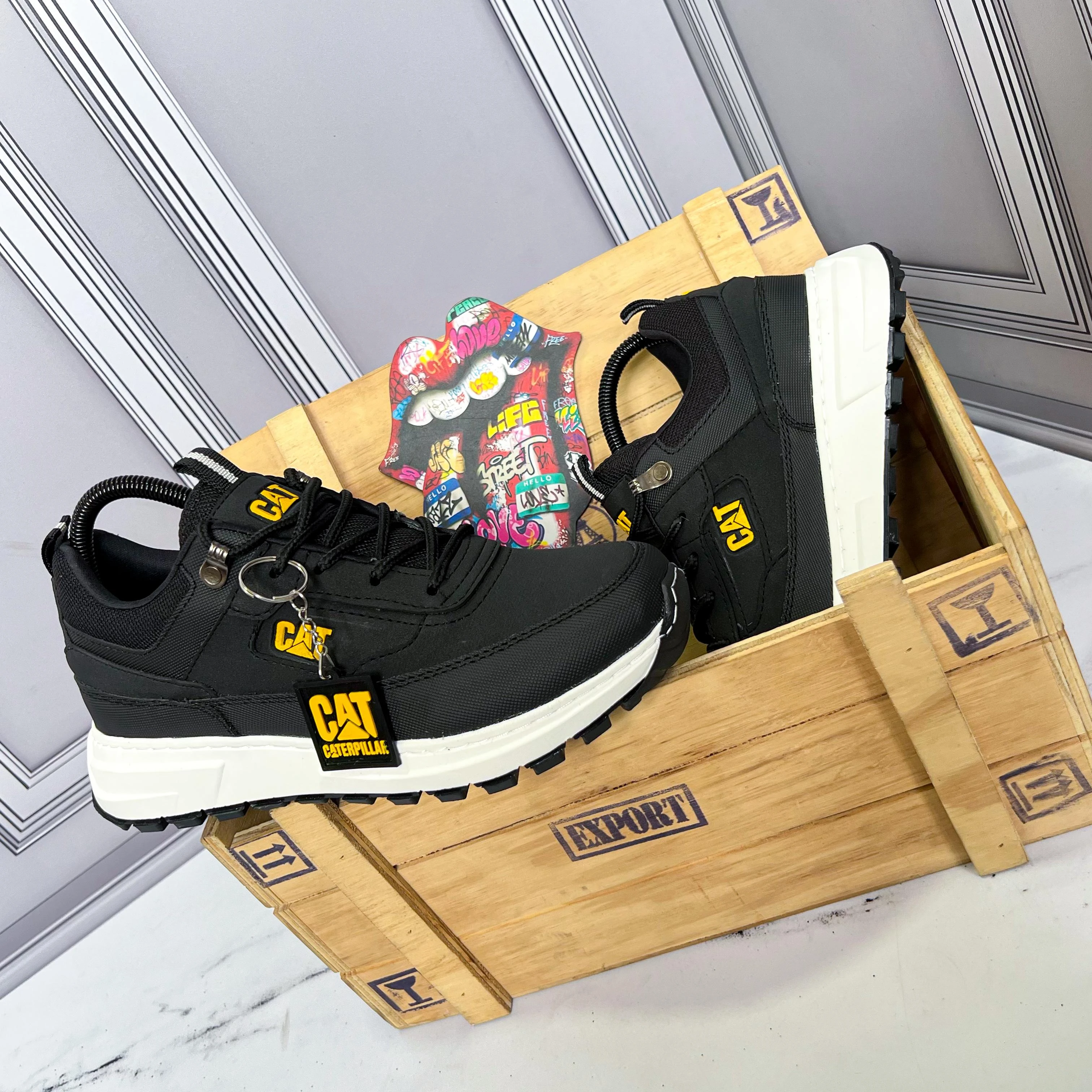 Zapato deportivo Caterpillar negro con suela blanca, repujado en amarillo y etiqueta colgante, presentado en caja de madera junto a bolsa de regalo.