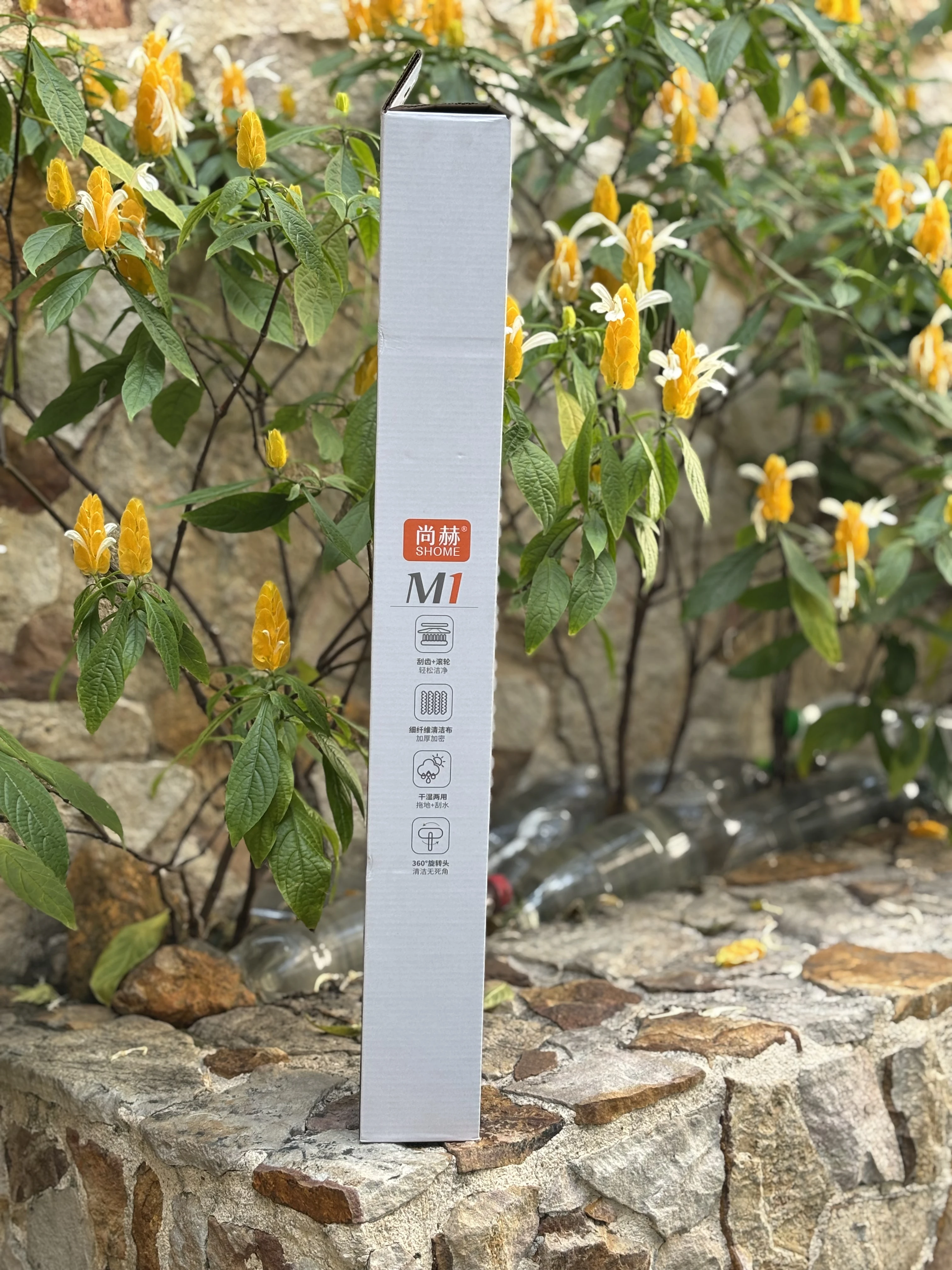 Trapeador multiusos M1 en caja blanca vertical, con logotipo rojo y detalles gráficos, colocado sobre muro de piedra con plantas de flores amarillas al fondo.