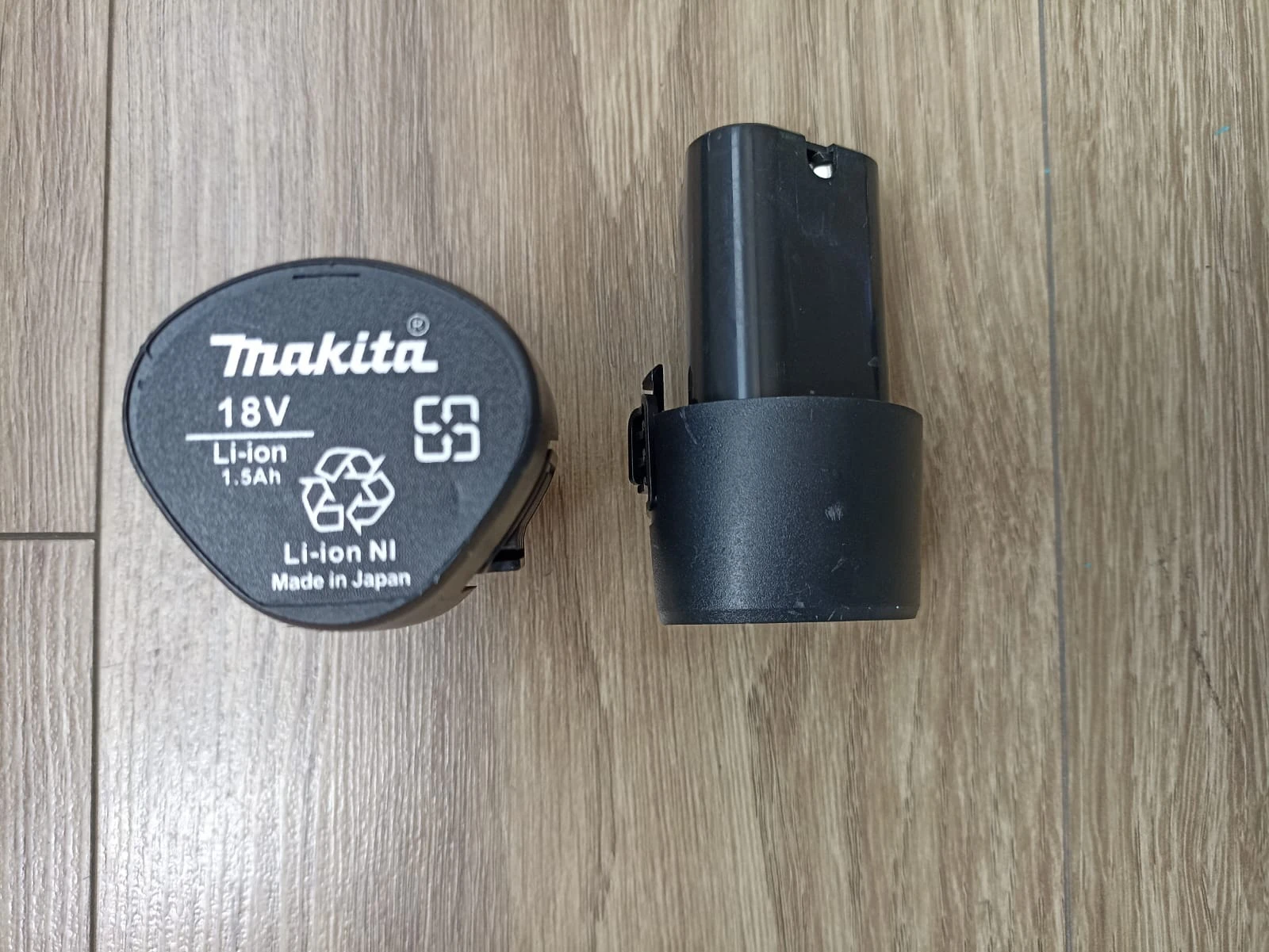 Batería Makita 18V Li-ion de 1.5Ah con etiqueta de reciclaje y número de serie, junto a su base de carga negra sobre superficie de madera.