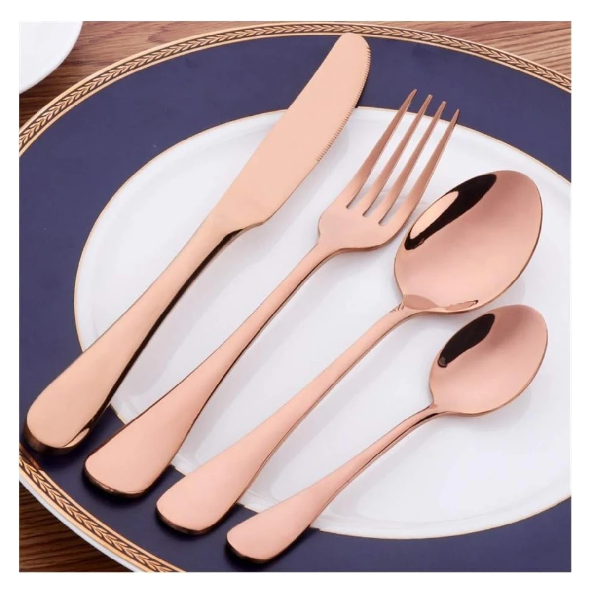 Set de cubiertos en acero inoxidable con acabado dorado rosa, mostrando cuchillo, tenedor y dos cucharas sobre plato blanco con borde azul y diseño dorado.