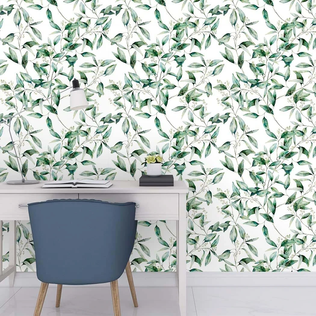 Papel adhesivo decorativo mate con patrón de hojas verdes sobre fondo blanco, aplicado en pared trasera de escritorio moderno con silla azul y accesorios minimalistas.