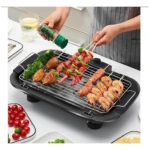 Parrillera eléctrica compacta en uso, con brochetas de carne y mariscos sobre rejilla, acompañada de vegetales y salsas en mesa de cocina.