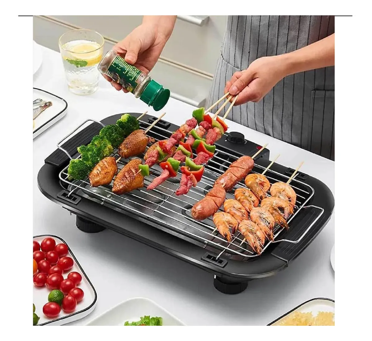 Parrillera eléctrica compacta en uso, con brochetas de carne y mariscos sobre rejilla, acompañada de vegetales y salsas en mesa de cocina.