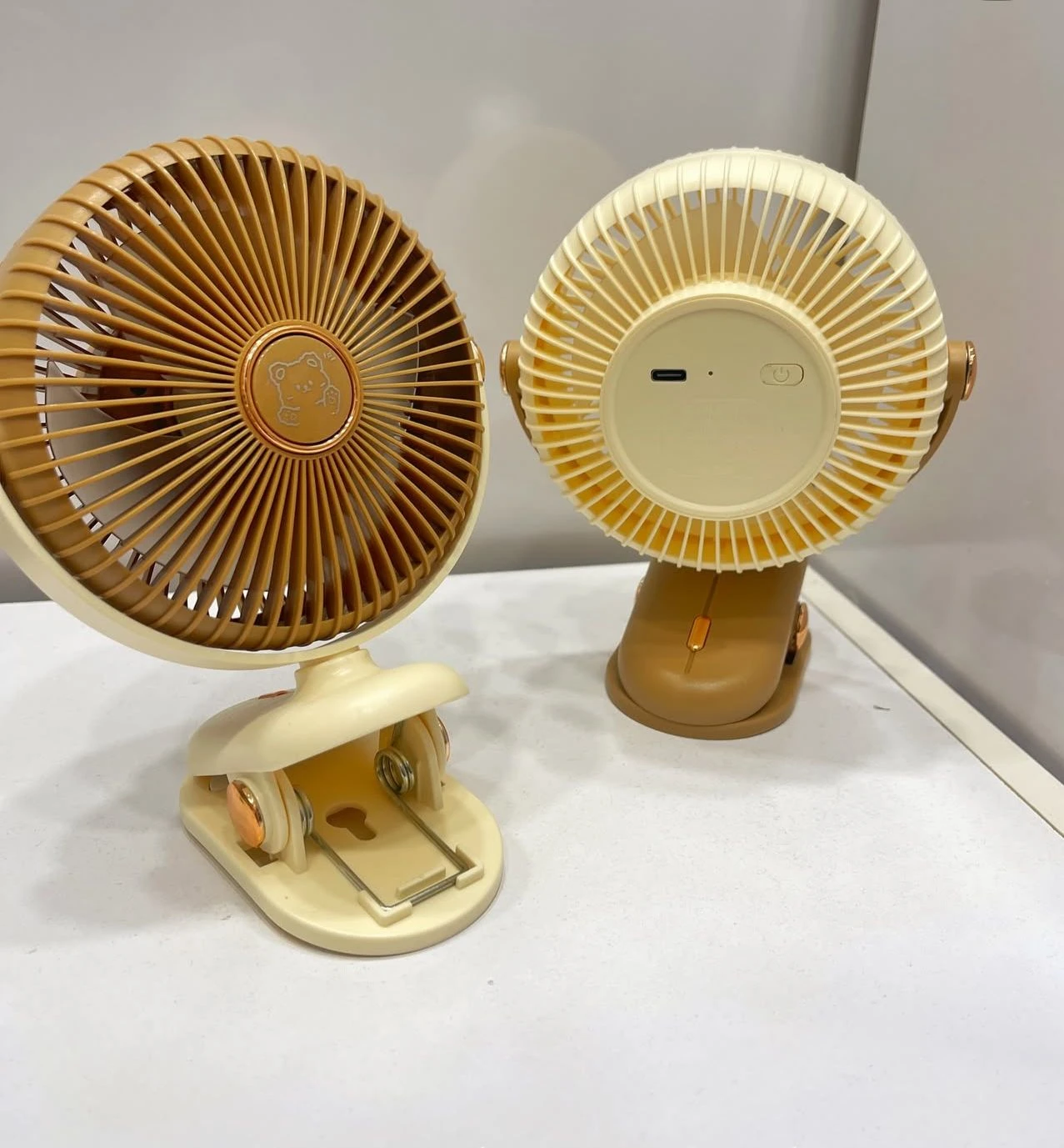 Dos ventiladores mini portátiles en tonos beige y marrón, con rejillas circulares y base de sujeción tipo pinza, mostrando frontal y trasera.