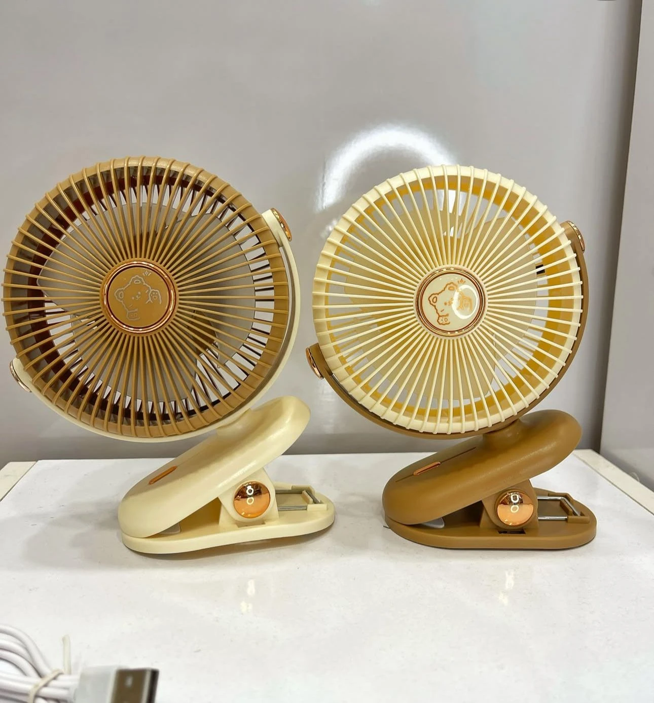 Dos ventiladores mini portátiles de color beige y marrón, con diseño circular y base de sujeción tipo pinza, dispuestos sobre superficie blanca.