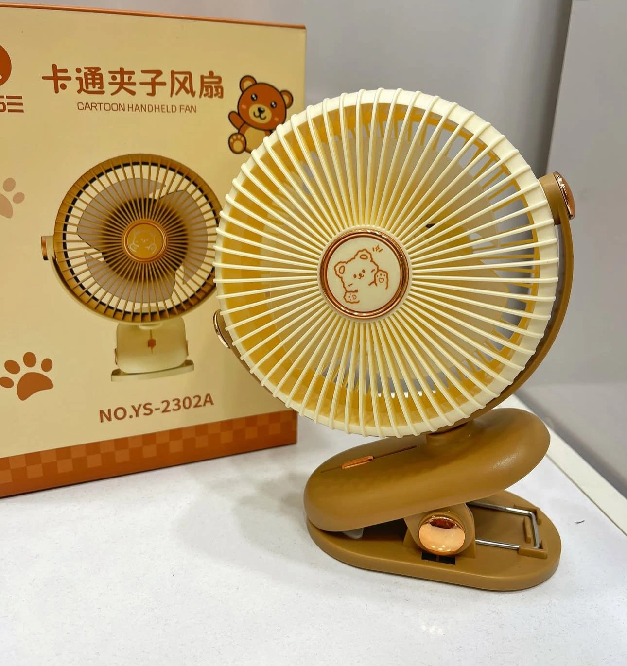 Ventilador mini portátil color beige con diseño de oso, montado en pinza para superficies. Incluye caja original con del producto y modelo NO.YS-2302A.