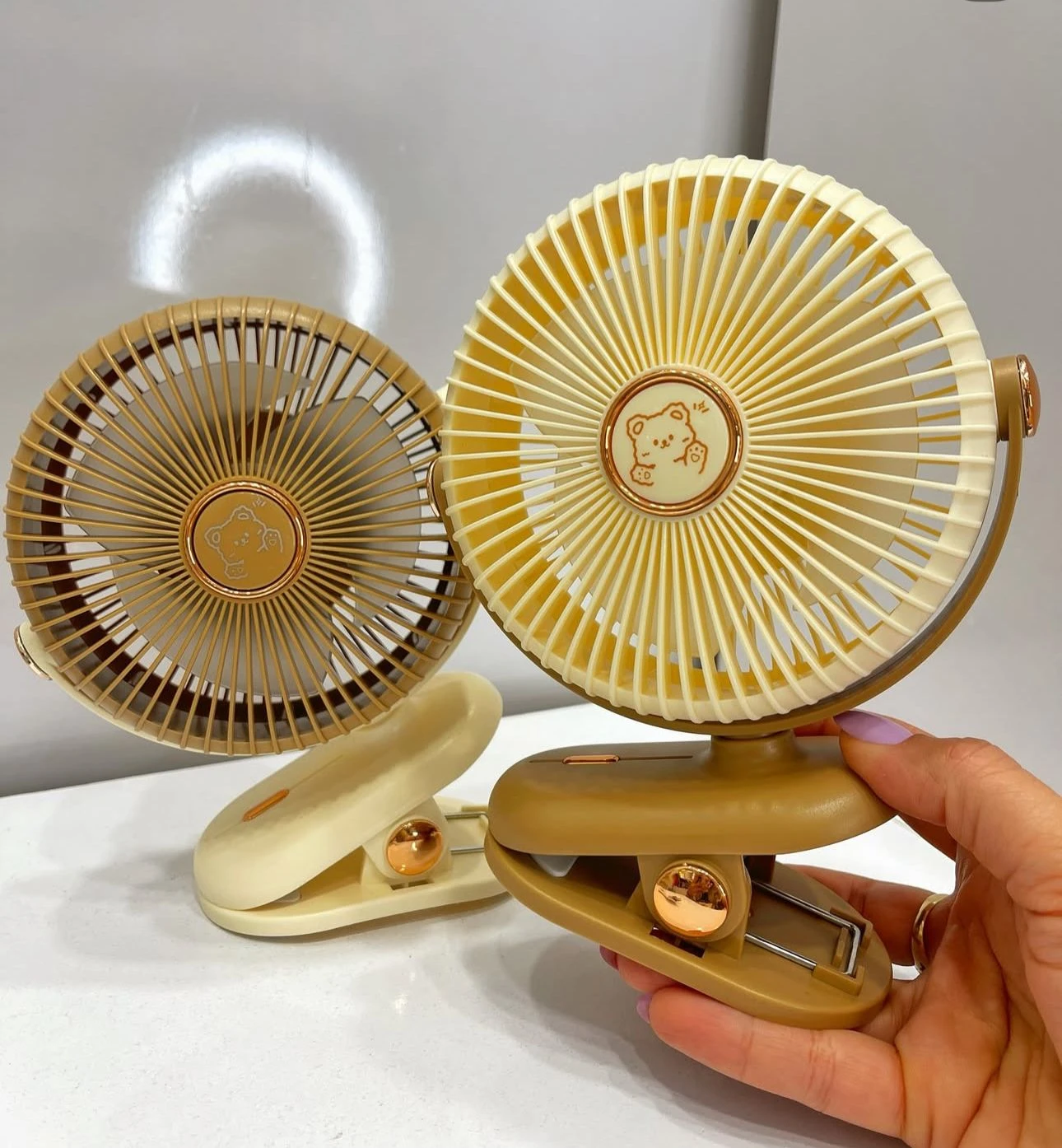Dos ventiladores mini portátiles con clip, uno en tono beige y otro en marrón claro, mostrando su diseño circular con rejilla protectora y logotipo central.