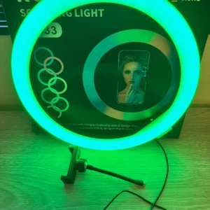 Aro de luz RGB de 33 cm en color verde brillante, montado sobre trípode ajustable, con cable visible y caja de producto al fondo.