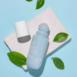 Desodorante roll-on Amway en tono azul claro con tapa blanca, sobre superficie texturizada beige, rodeado de hojas verdes. Presentación limpia y minimalista.