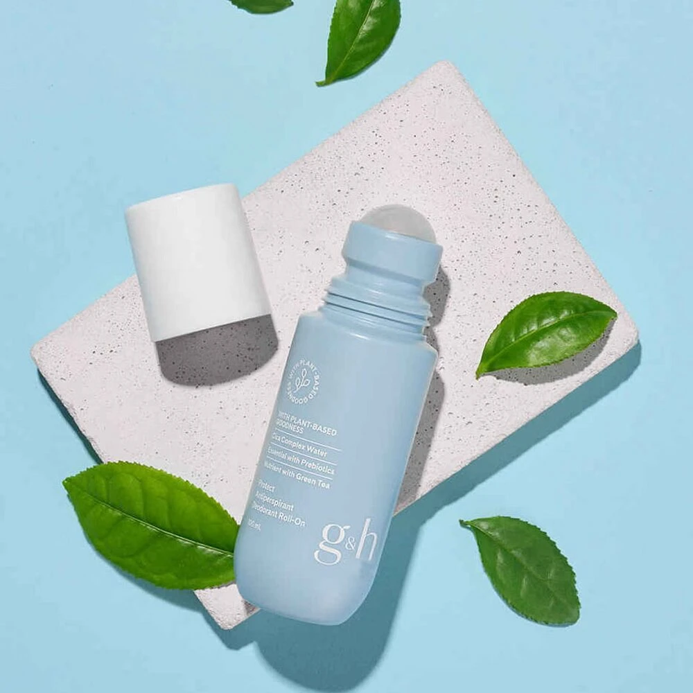 Desodorante roll-on Amway en tono azul claro con tapa blanca, sobre superficie texturizada beige, rodeado de hojas verdes. Presentación limpia y minimalista.