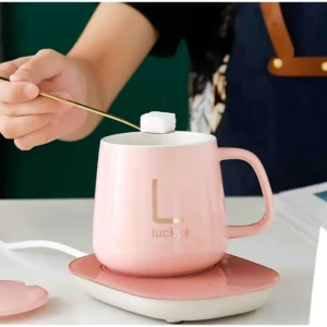 Termo eléctrico de cerámica rosa con base calefactora, en uso para calentar café o té, con logo dorado y tapa removible visible.