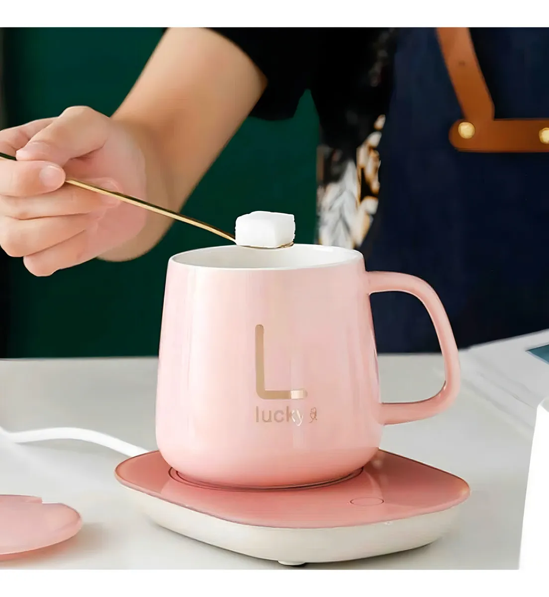 Termo eléctrico de cerámica rosa con base calefactora, en uso para calentar café o té, con logo dorado y tapa removible visible.