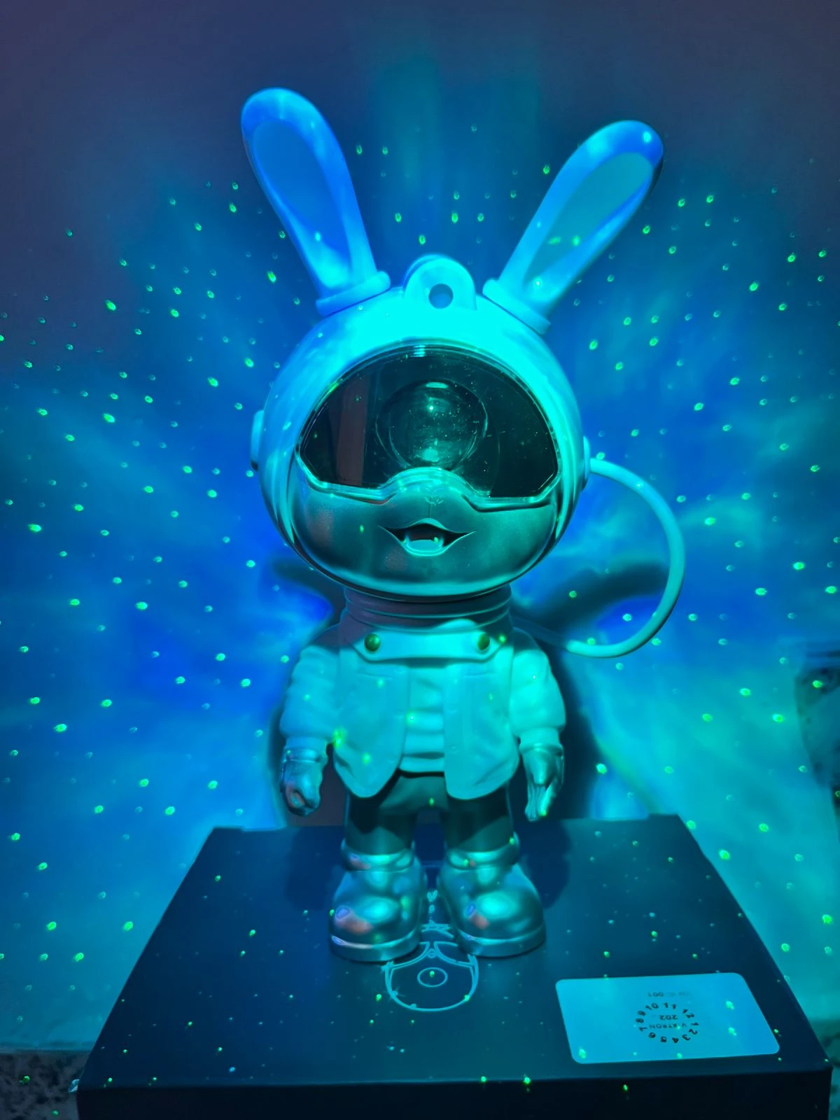 Proyector de galaxias en forma de conejo astronauta, color plateado con detalles azules, emitiendo luz estelar verde sobre superficie oscura.