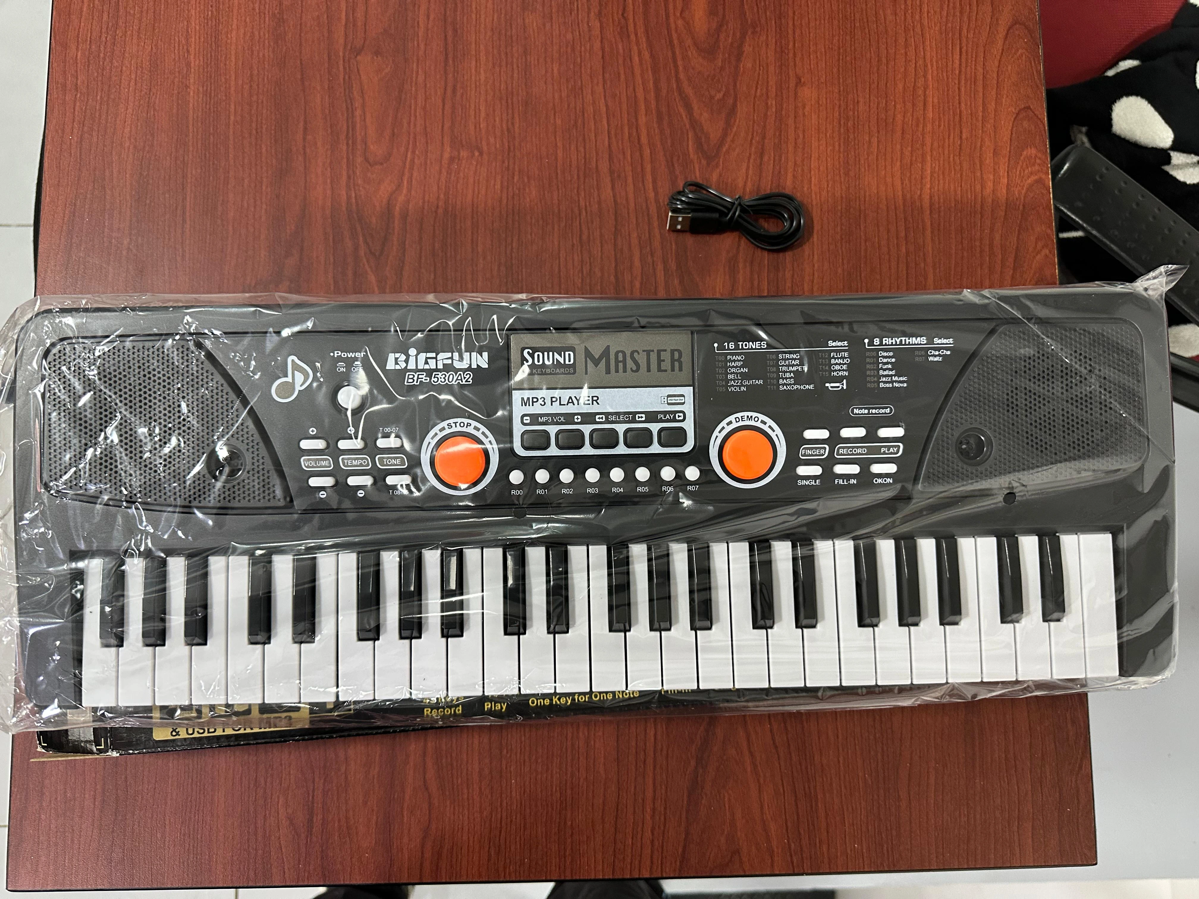 Teclado electrónico de 61 teclas negro con pantalla digital y dos botones naranjas, envuelto en plástico transparente sobre superficie de madera.