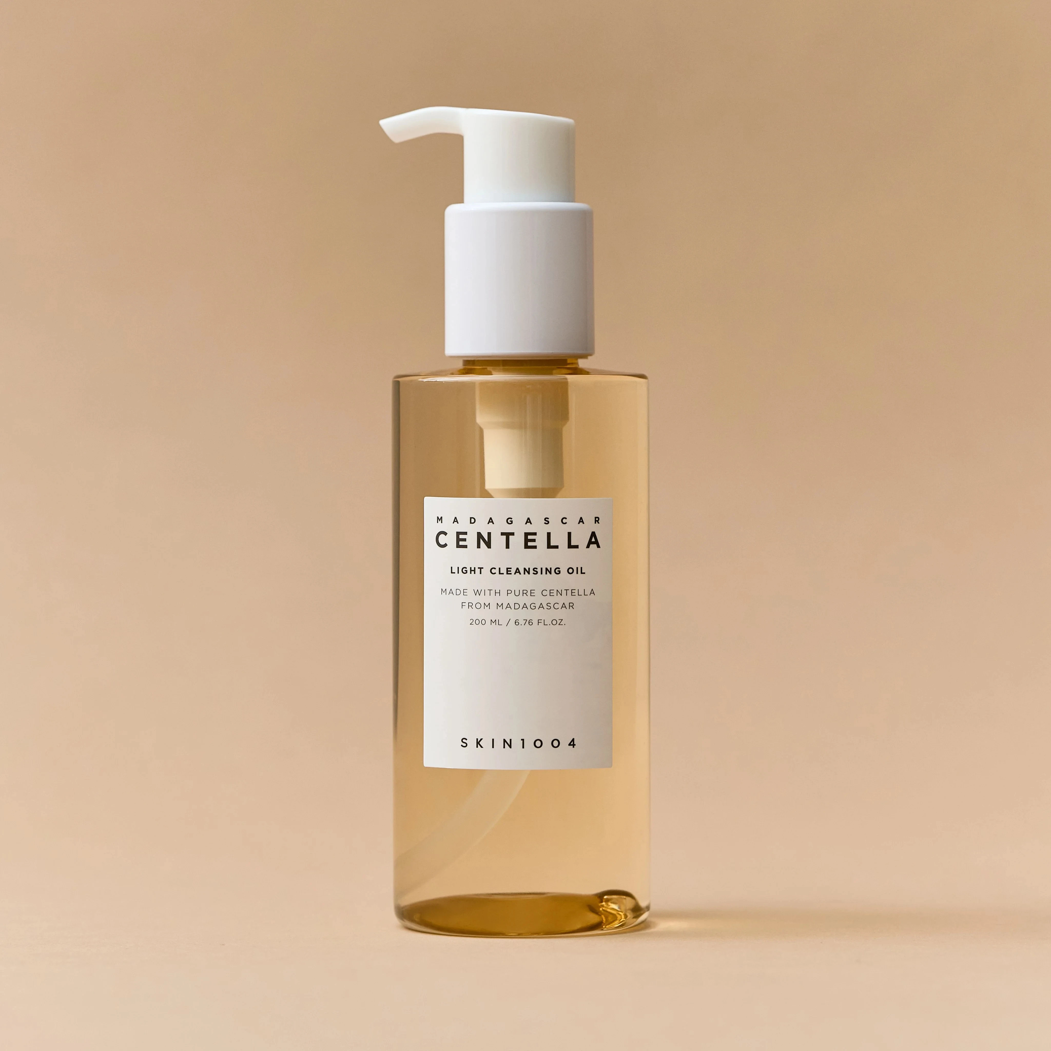 Botella transparente con aceite limpiador facial de color ámbar, etiqueta blanca que indica Centella Asiática de Madagascar, dispensador blanco y fondo beige neutro.