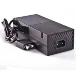 Fuente de alimentación externa negra para Xbox One, con cable enrollado y conector C14 visible en la parte trasera.
