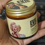 Contenedor de crema Evil Goods con tapa dorada y etiqueta amarilla que muestra retratos de Marilyn Monroe y un hombre, sobre teclado.