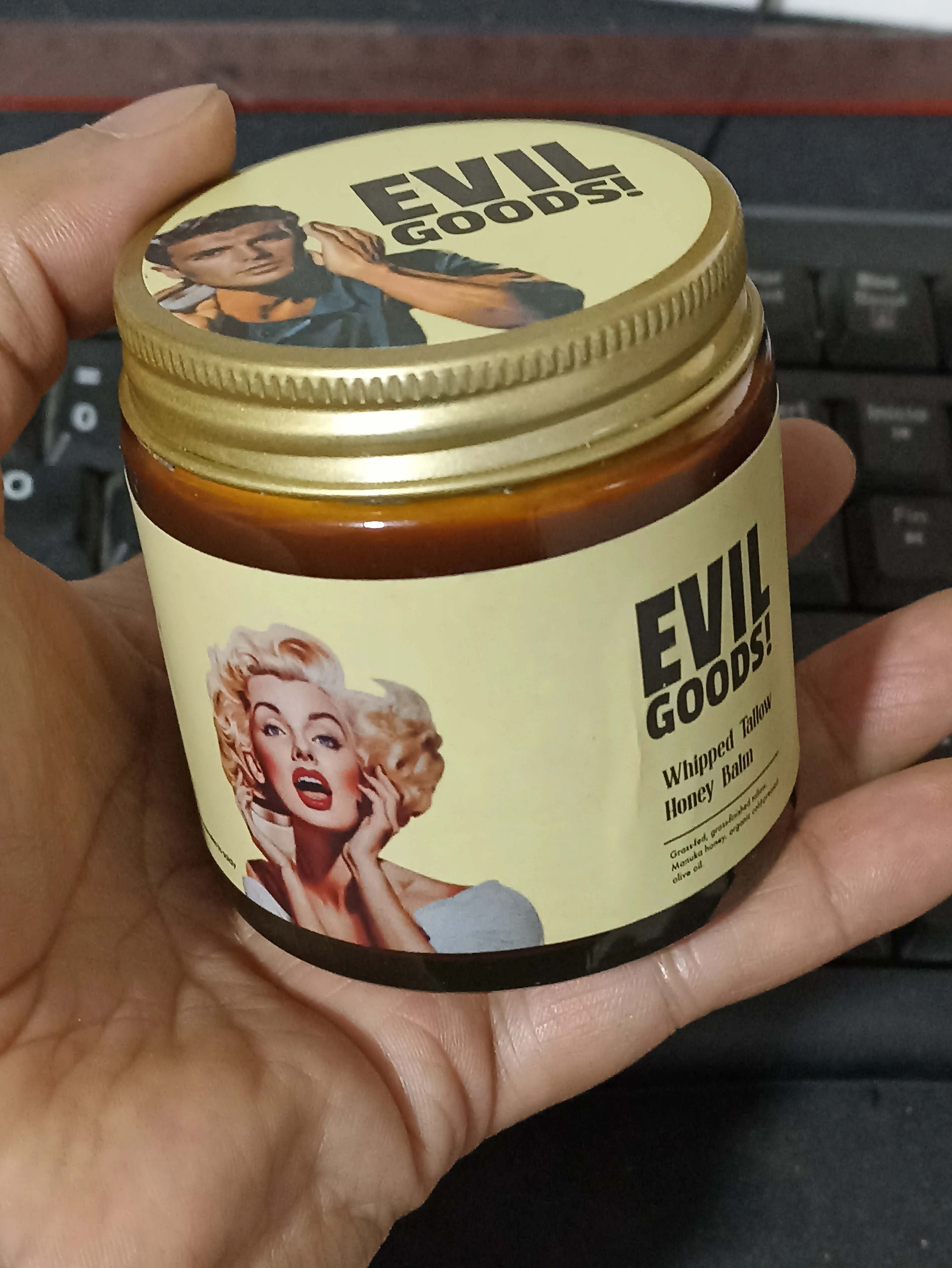 Contenedor de crema Evil Goods con tapa dorada y etiqueta amarilla que muestra retratos de Marilyn Monroe y un hombre, sobre teclado.