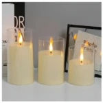 Tres velas LED sin llama en recipientes de vidrio transparente, de diferentes alturas, encendidas sobre superficie blanca, con marco decorativo y libros al fondo.