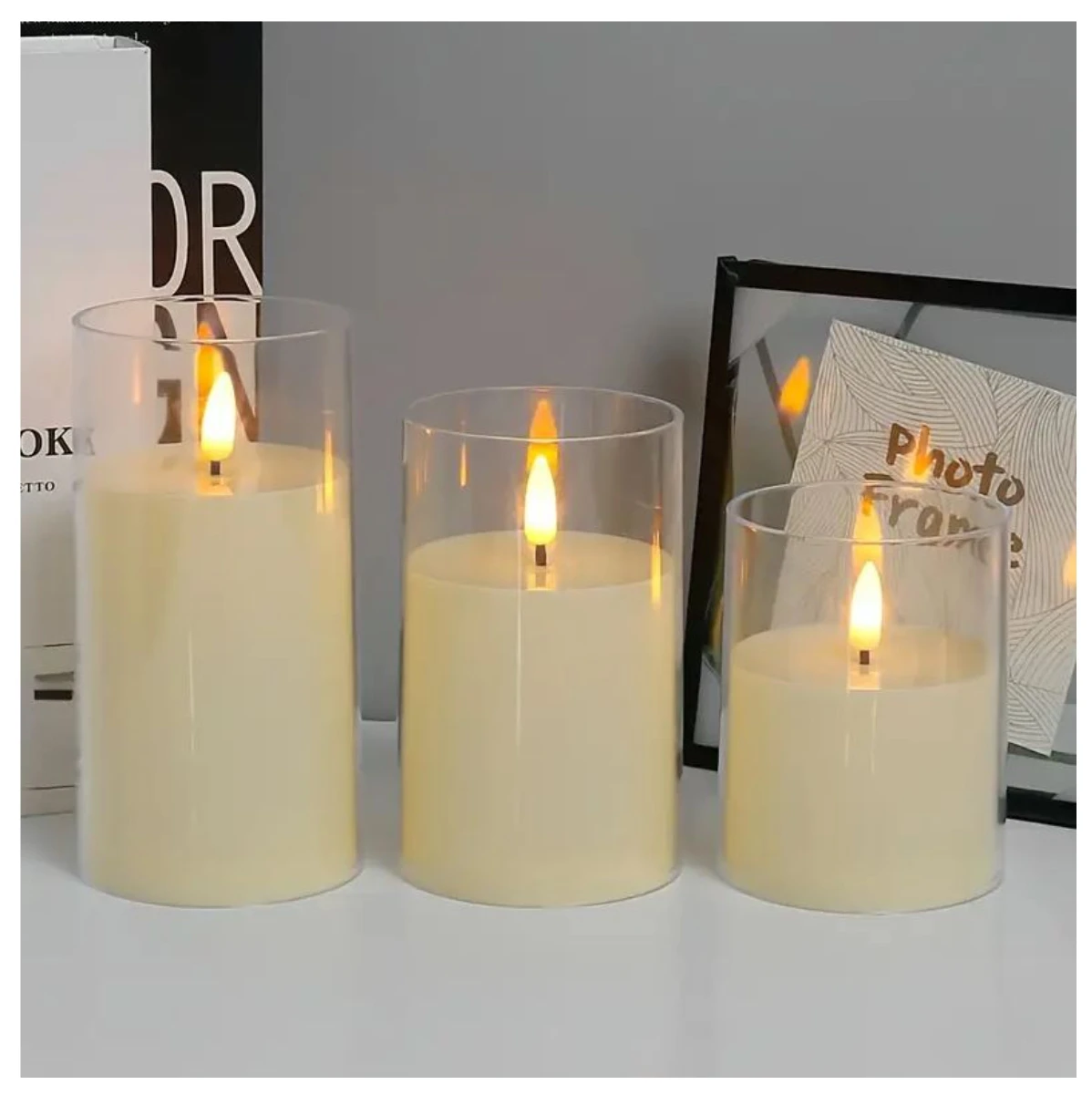 Tres velas LED sin llama en recipientes de vidrio transparente, de diferentes alturas, encendidas sobre superficie blanca, con marco decorativo y libros al fondo.