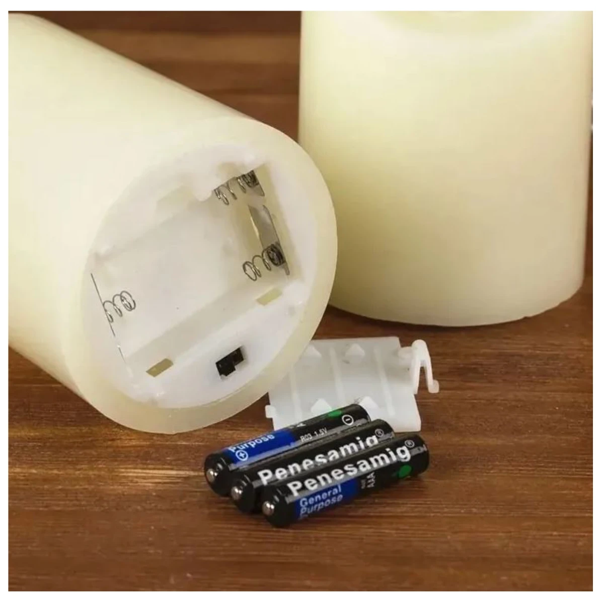 Vela LED sin llama de color crema, mostrando su base con compartimento para pilas AA y dos baterías incluidas, junto a otra vela encendida en segundo plano sobre superficie de madera.