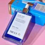 Bote transparente de aceite corporal Glass Skin de Truly, con etiqueta blanca y detalles dorados, sobre fondo rosa. Incluye caja azul con nombre del producto visible.