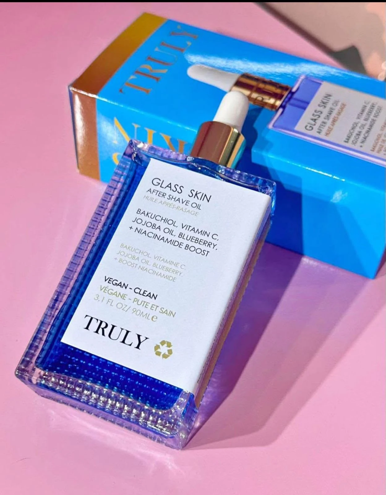 Bote transparente de aceite corporal Glass Skin de Truly, con etiqueta blanca y detalles dorados, sobre fondo rosa. Incluye caja azul con nombre del producto visible.