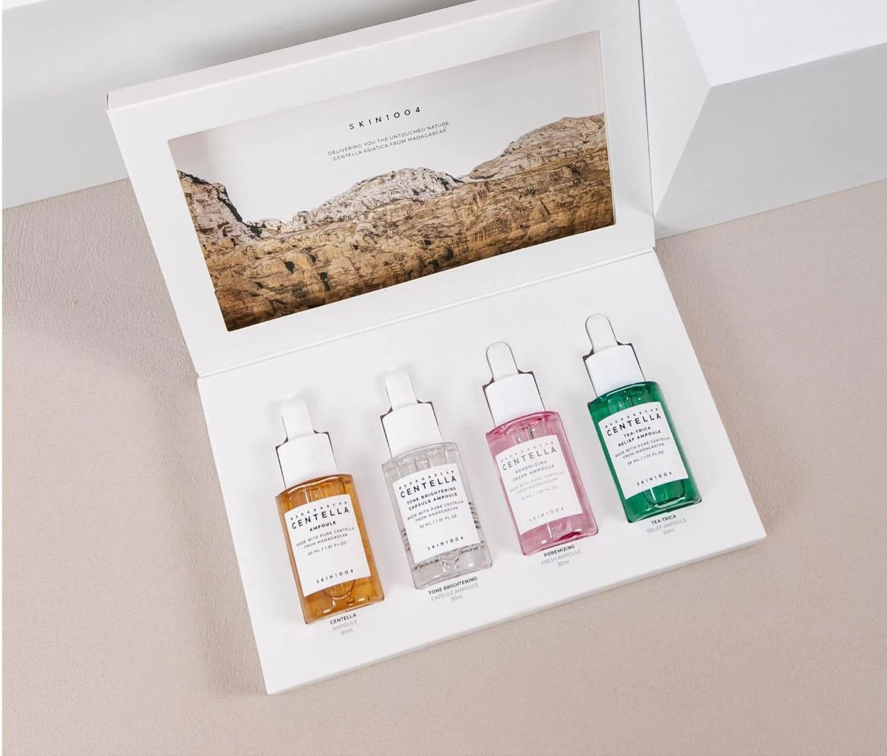 Kit de cuatro mini ampollas SKIN1004 con centella asiática, presentadas en caja blanca con tapa ilustrada. Frascos transparentes con líquidos en tonos amarillo, blanco, rosa y verde, dispuestos horizontalmente sobre fondo claro.