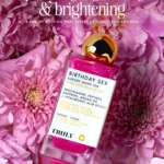 Botella de aceite corporal Truly Birthday Sex con brillo rosa, sobre flores rosadas. Etiqueta blanca con ingredientes visibles: niacinamida, péptidos, argán y ácido hialurónico. Presentación vegana y limpia.