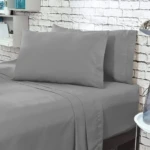 Juego de sábanas gris claro queen liso, con dos almohadas y sábanas ajustadas, sobre cama en habitación con pared de ladrillo blanco.