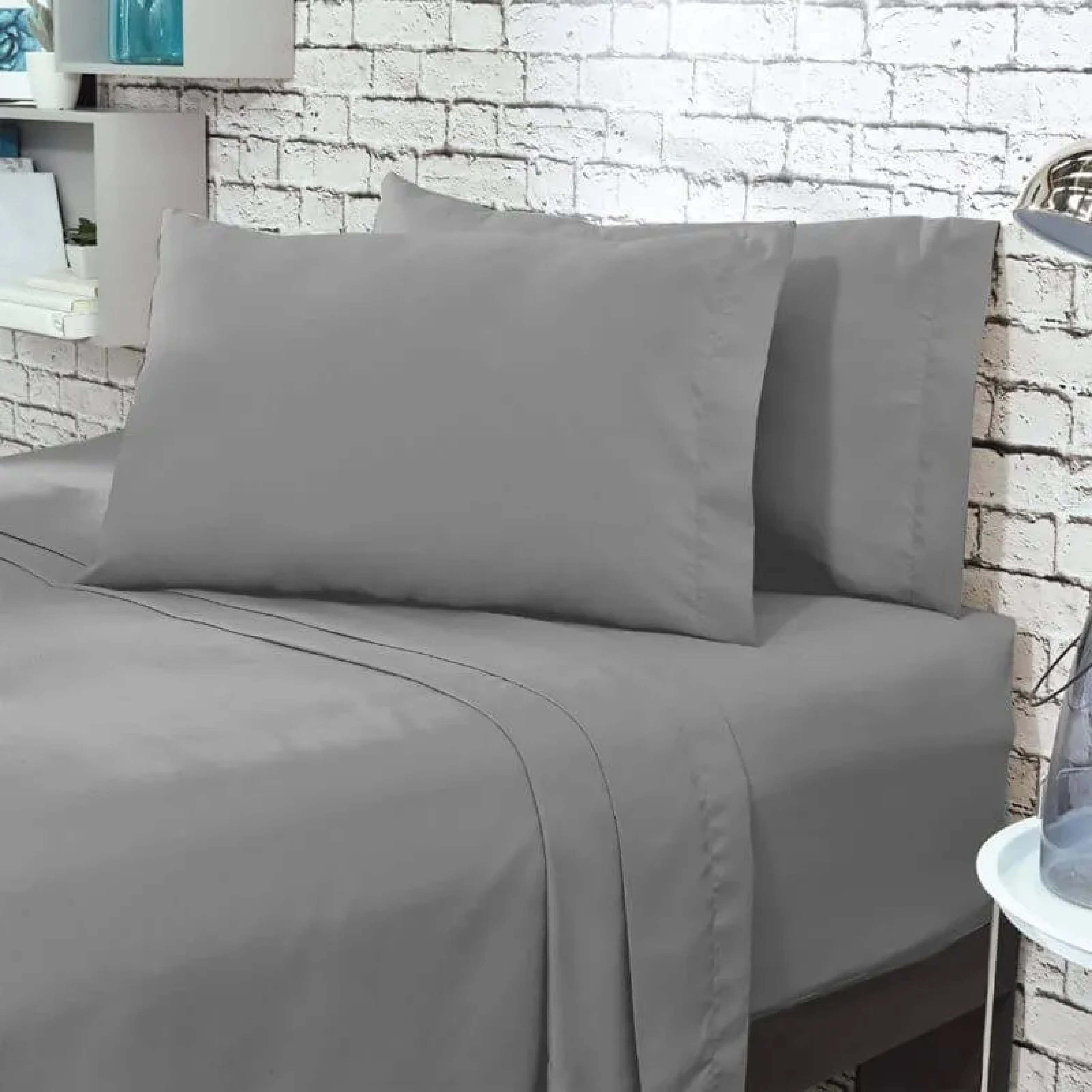 Juego de sábanas gris claro queen liso, con dos almohadas y sábanas ajustadas, sobre cama en habitación con pared de ladrillo blanco.