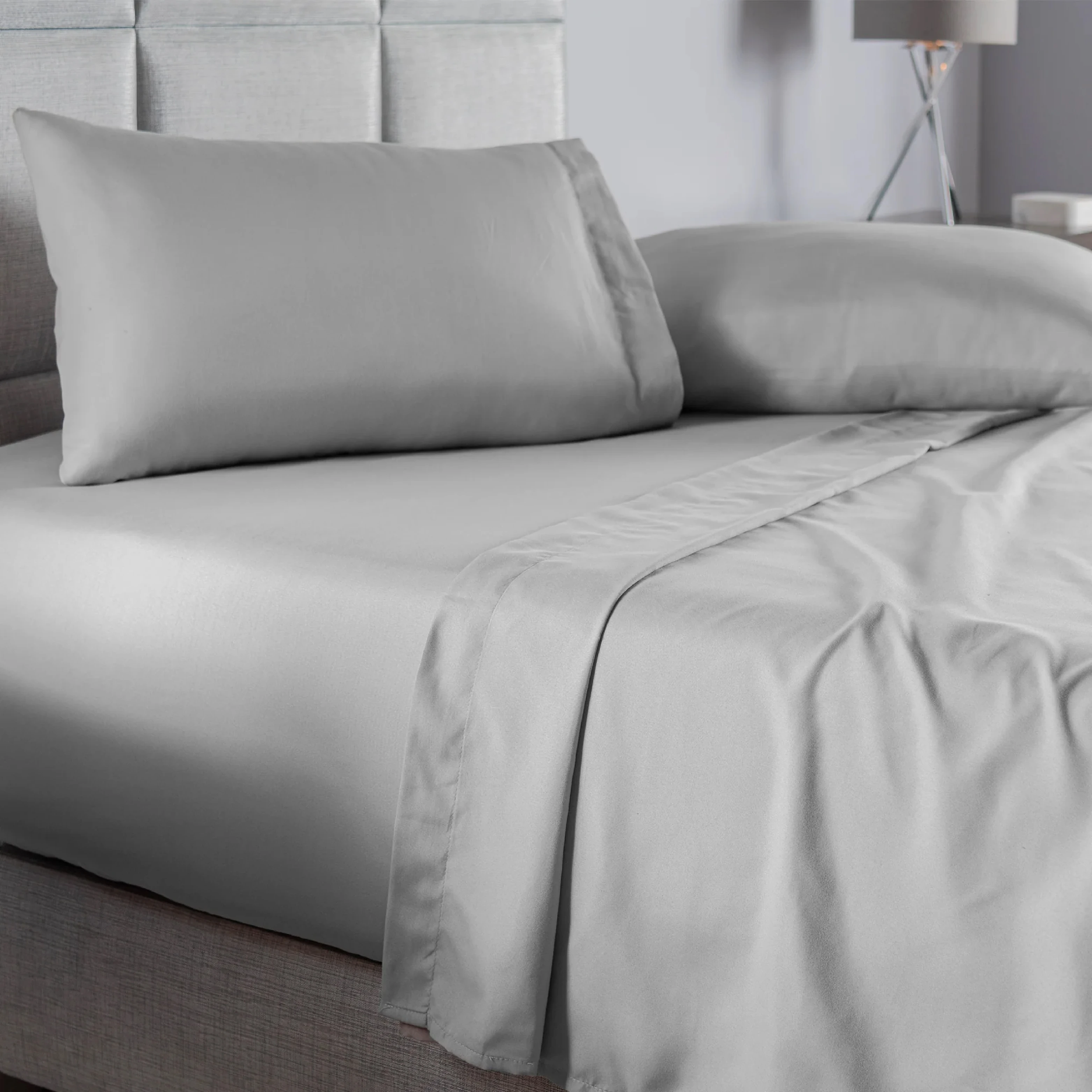 Juego de sábanas gris claro queen liso, con acabado suave y moderno, mostrando colchón, almohadas y sábanas dobladas en cama con cabecero tapizado.