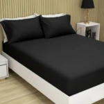 Sábana ajustable negra con dos fundas de almohada, vista frontal en cama blanca sobre suelo de madera clara.