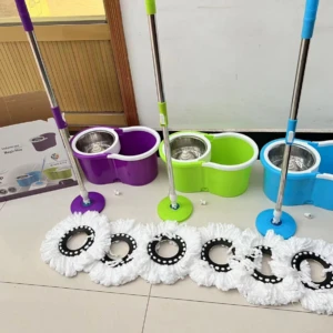 Set de traperos mágicos con cabezal giratorio y escurrido automático, en colores morado, verde y azul, con sus respectivos mangos y cabezales blancos dispuestos sobre suelo de baldosas.