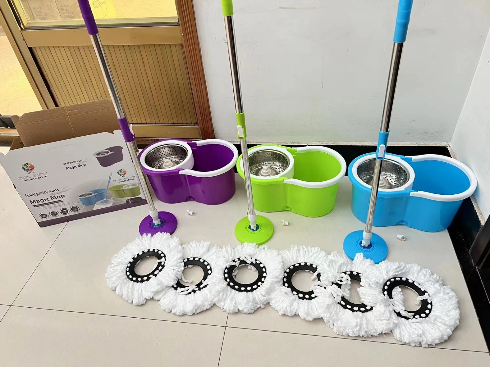 Set de traperos mágicos con cabezal giratorio y escurrido automático, en colores morado, verde y azul, con sus respectivos mangos y cabezales blancos dispuestos sobre suelo de baldosas.