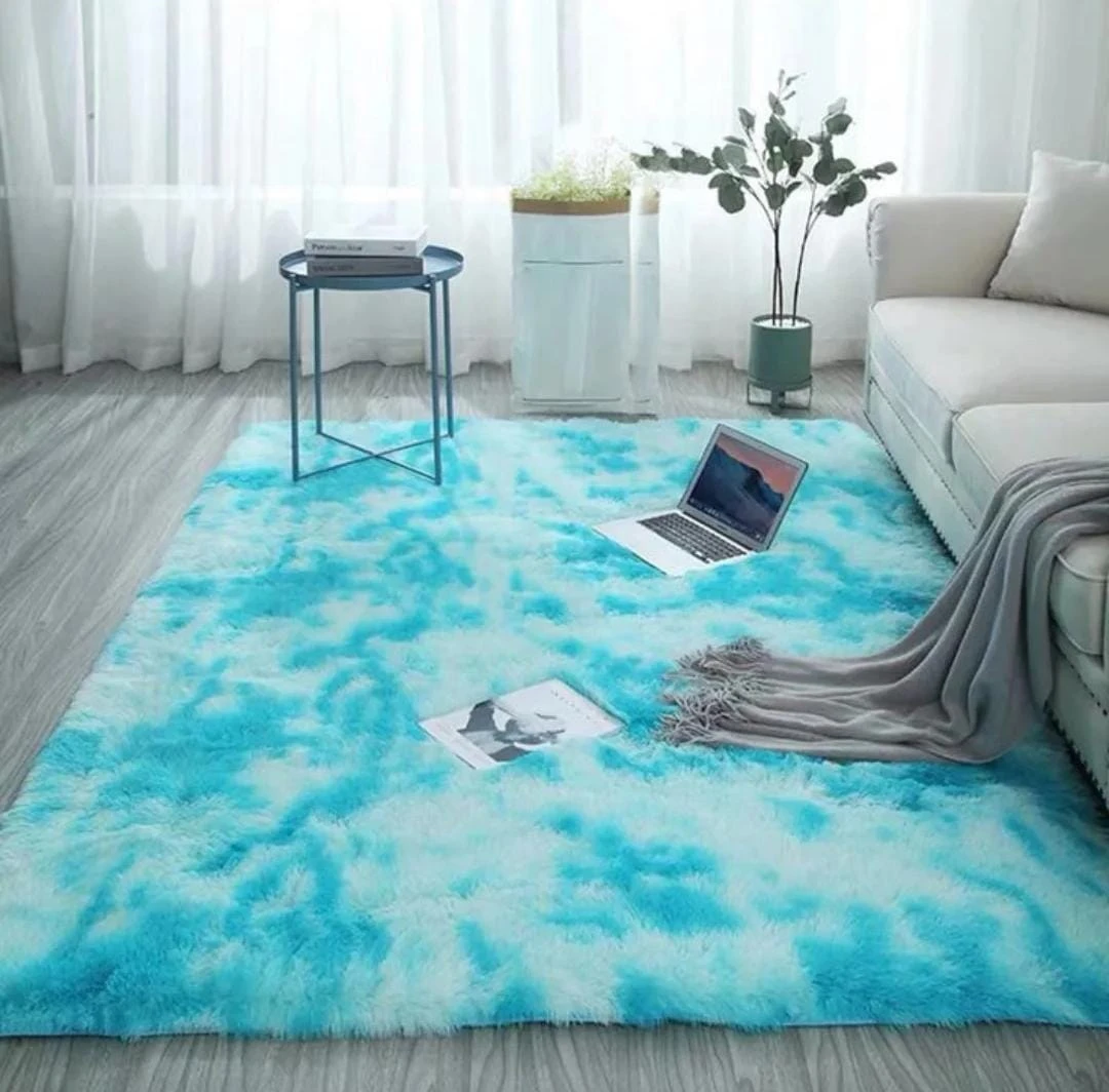 Tapete peludo grande de alta densidad en tonos azul cielo y blanco, con textura suave y larga, colocado en sala junto a sofá, laptop y mesa auxiliar.