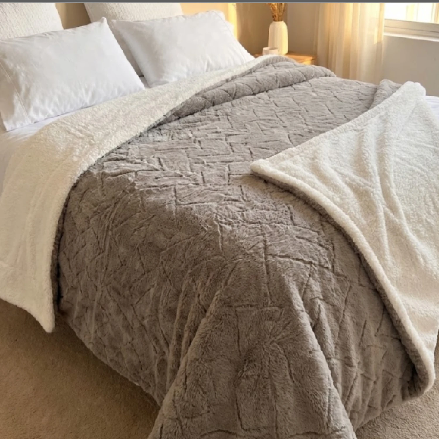 Cobija gris de doble cara con textura de ovejero y franela, mostrando su parte superior acolchada y la reversible blanca en cama con sábanas blancas.