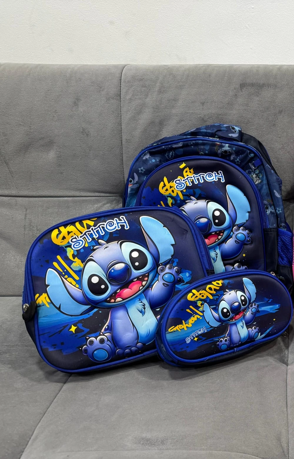 Morral escolar azul con estampado de Stitch, acompañado de lonchera y estuche del mismo diseño, sobre sofá gris.