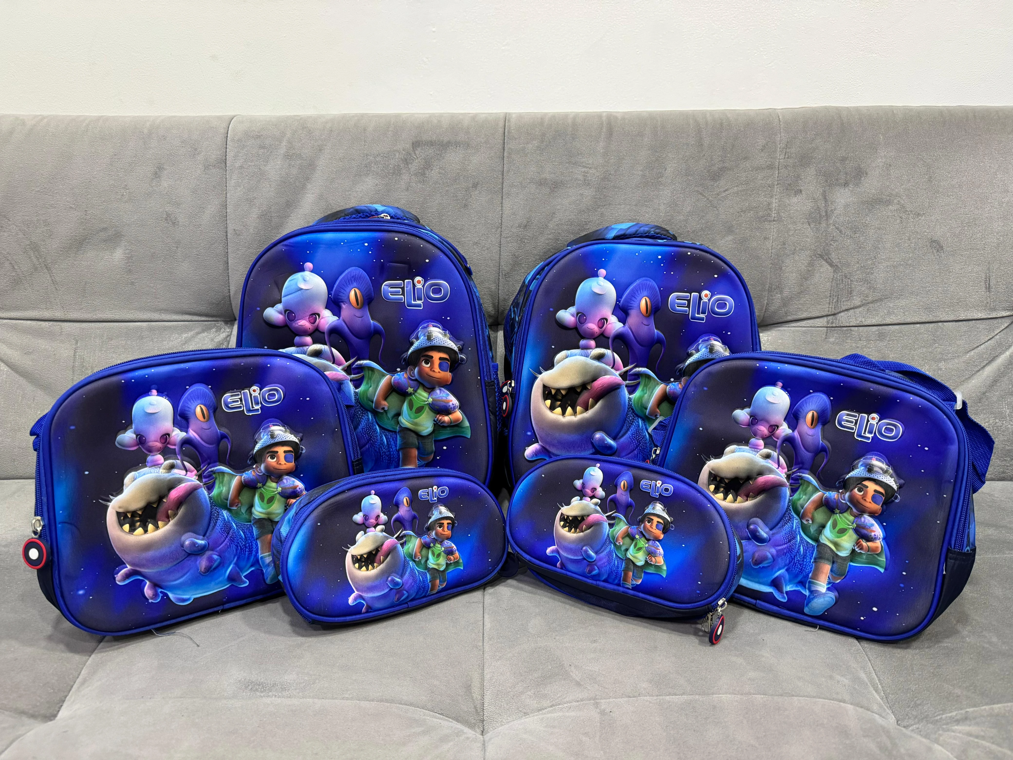 Set de mochila escolar azul con personajes 3D, lonchera y estuche para útiles, sobre sofá gris.
